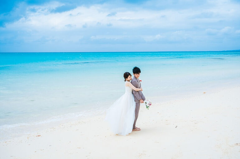 青いビーチの前で新婦が後ろから新郎をハグする様子